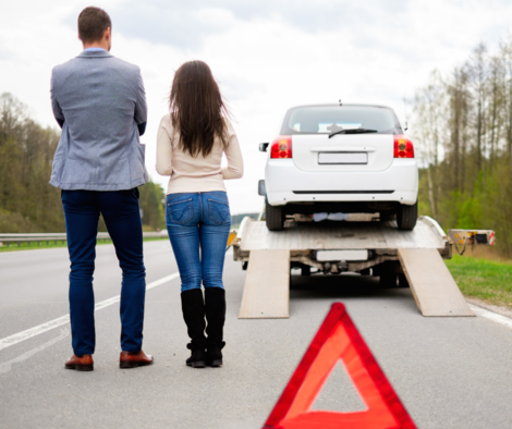 Man and women watching their car leaving on a tow truck with a traffic warning triangle in the forreground